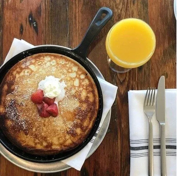 Overhead pancake in skillet with strawberries, cream, juice, and cutlery.