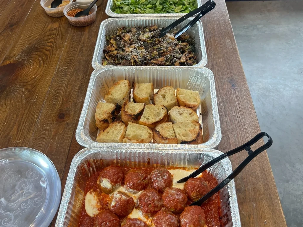 Foil trays of salad, roasted vegetables, and meatballs on wooden table.
