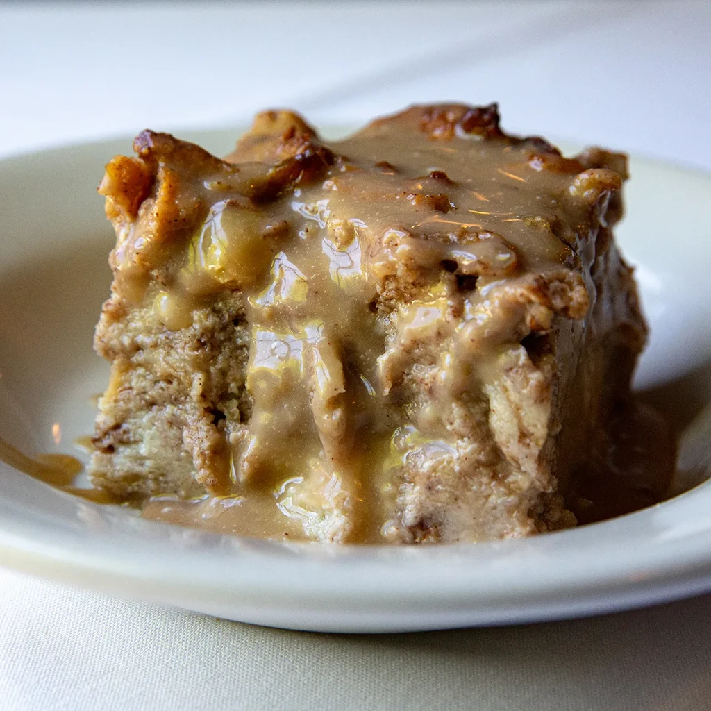 Square bread pudding slice with creamy caramel sauce on a cream plate.