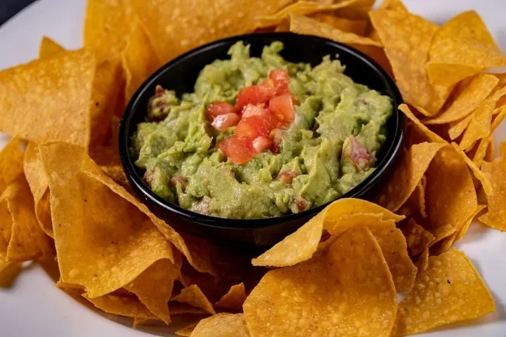 Black bowl of chunky guacamole with tomatoes, surrounded by tortilla chips.