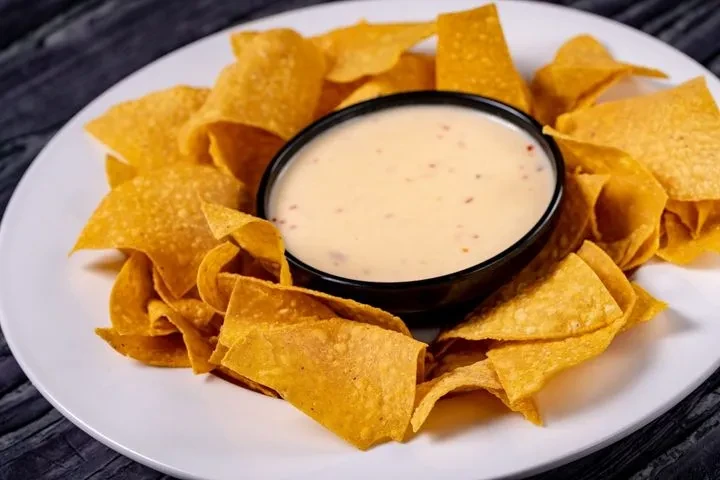 Golden tortilla chips around a bowl of yellow queso with red spice.