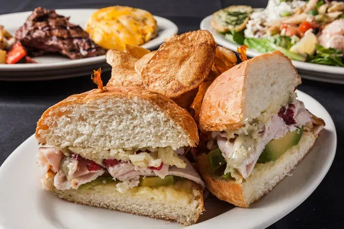 Plated sandwich with chips, blurred steak and salad in background.