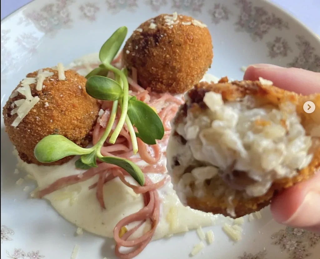 Hand holds broken fried arancini on sauce with microgreens and cheese.