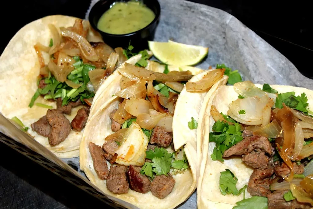 Three street-style steak tacos with onions, cilantro, lime, green sauce.