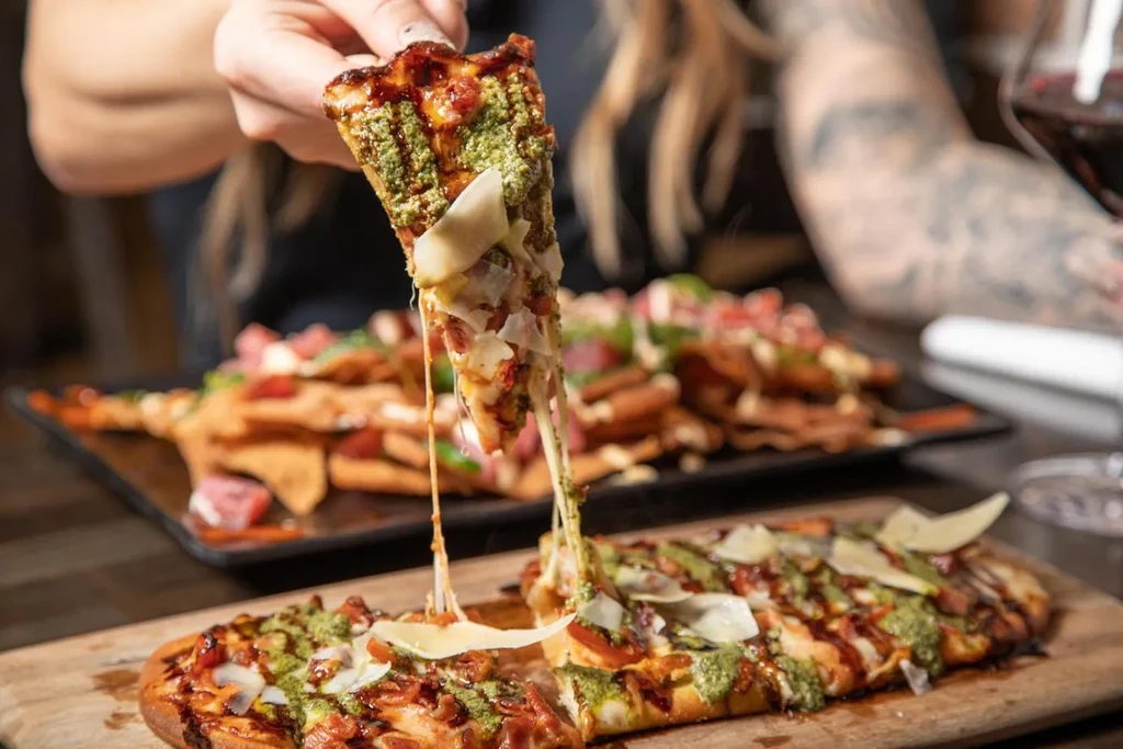 Hand lifts pesto flatbread with red sauce, showing a cheese pull.