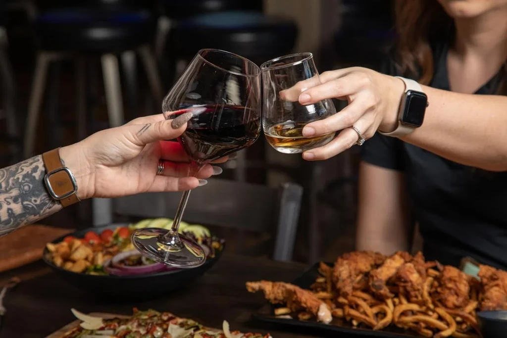 Hands clink red wine and amber spirit glasses over food.