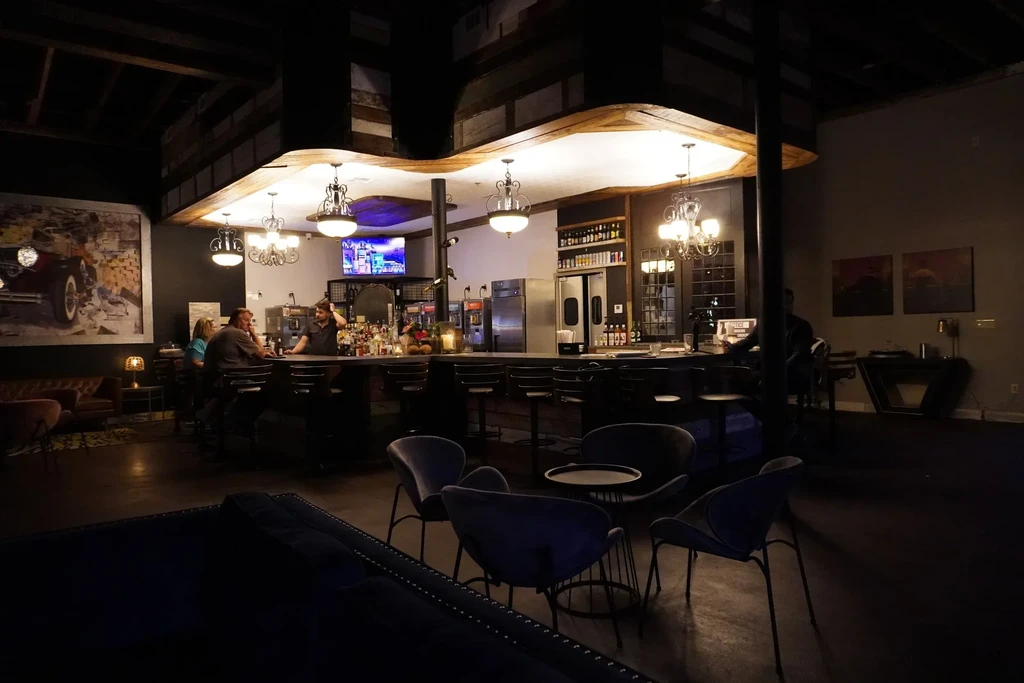 Dimly lit bar with bartender, patrons, wavy wooden ceiling, and lounge seating.