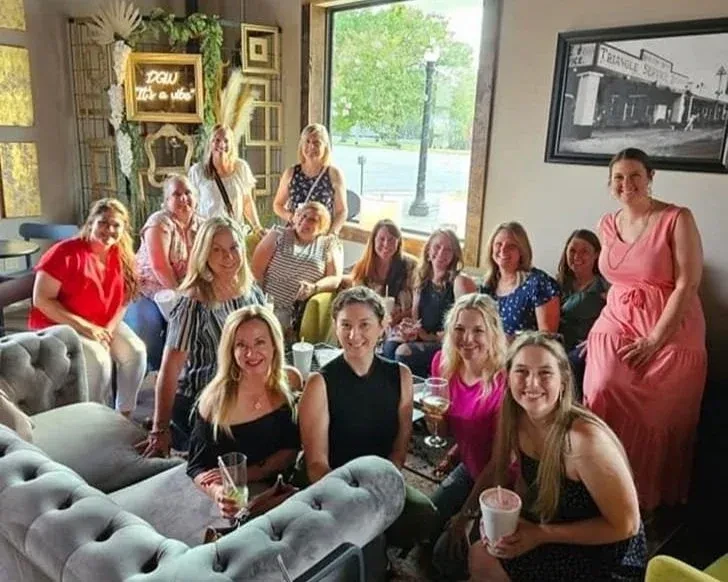 Smiling women gather in a sunlit cafe lounge with drinks.