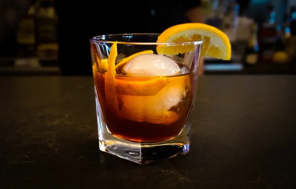 Amber cocktail in rocks glass with ice and orange garnish.