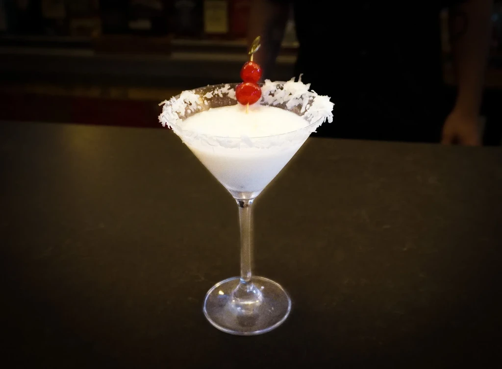 Creamy white cocktail in martini glass with coconut rim and cherry garnish.