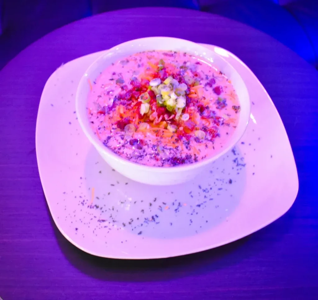 Pinkish soup with cheese, red bits, green onions, under purple lighting.
