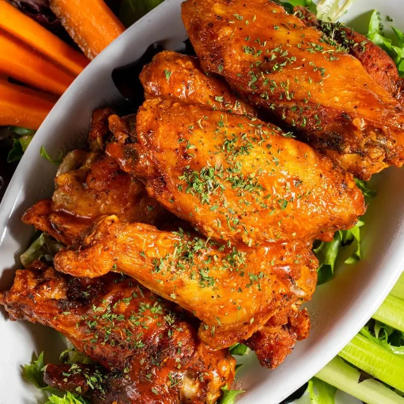 Saucy orange-red chicken wings with green herbs, carrot, and celery sticks.