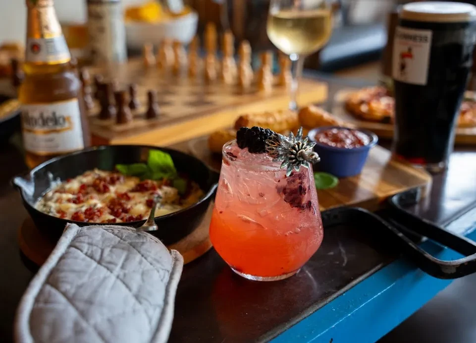 Cocktail, savory dish, beer, wine, and chessboard on a restaurant table.