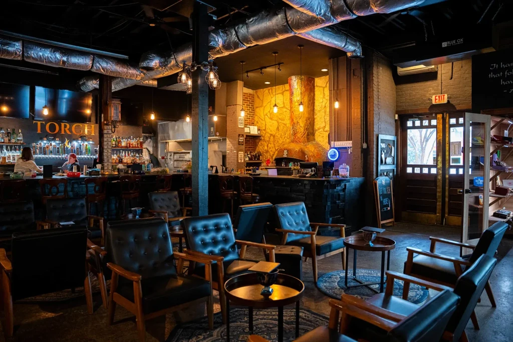 Dimly lit industrial restaurant with lounge chairs, bar, and pizza oven.