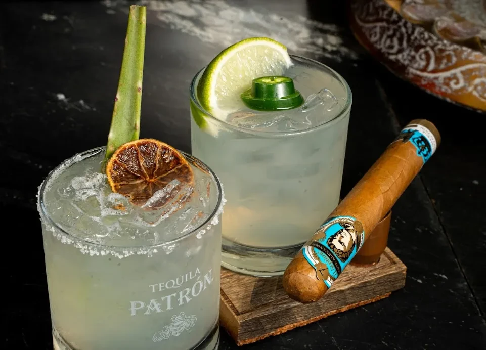 Two garnished cocktails and a banded cigar on a dark surface.