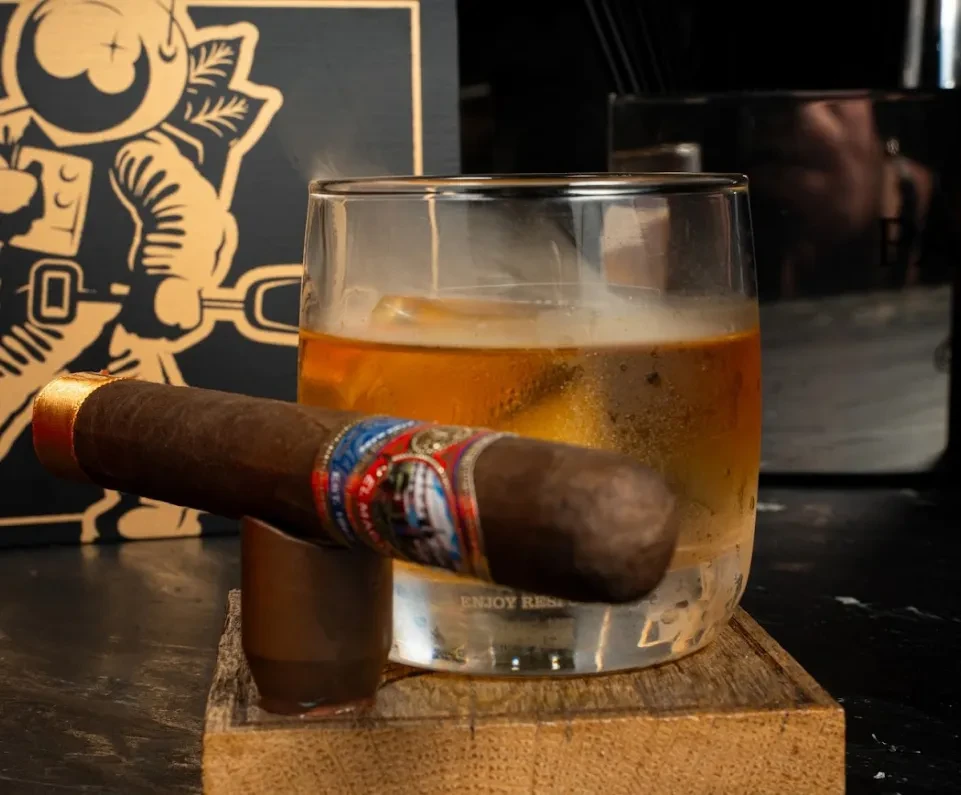 Whiskey on ice, a cigar, and a coaster in a dark bar.