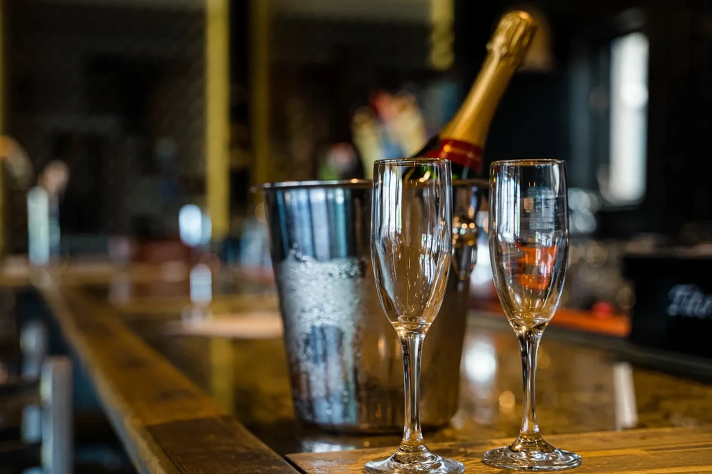 Two empty champagne flutes, bottle in ice bucket on wooden bar.