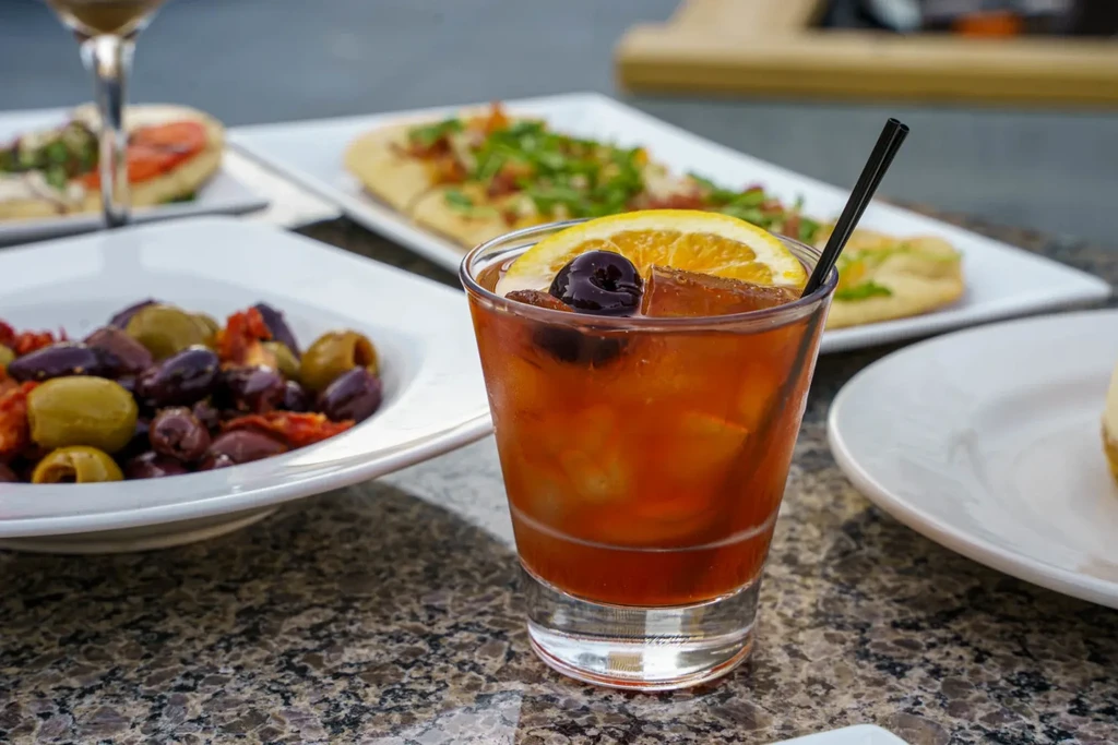 Rocks glass with amber cocktail, cherry, orange, and blurred food.