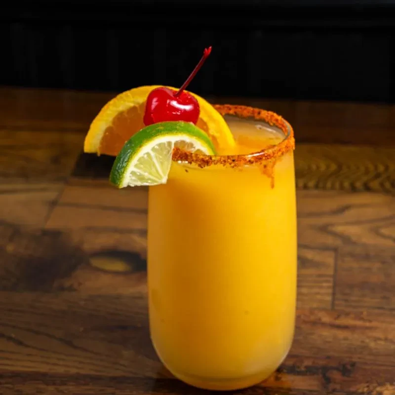 Yellow drink in clear glass, lime, orange, cherry, spiced rim.