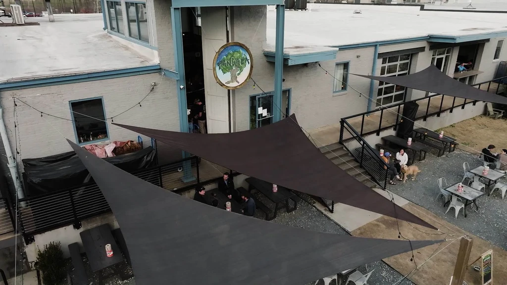 High-angle view: brewery patio with tree logo, shade sails, and patrons.