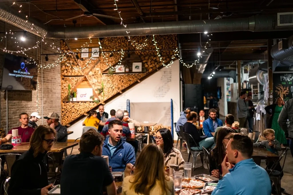 Diners eating pizza in an industrial-rustic restaurant with a firewood wall.