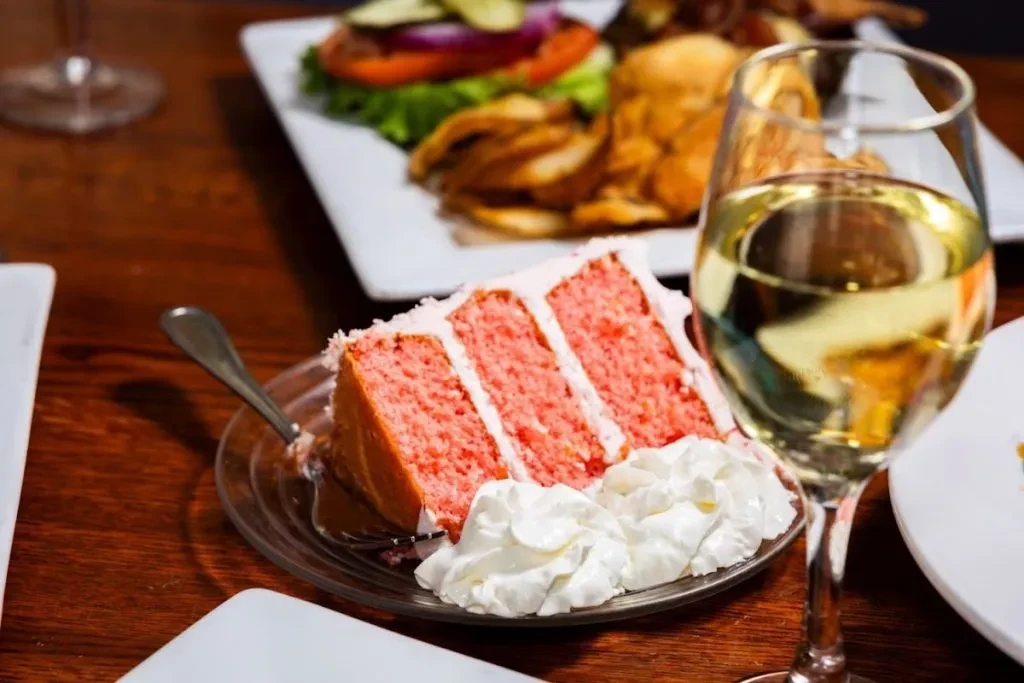 Pink layer cake, white wine, and burger meal on a dark wood table.