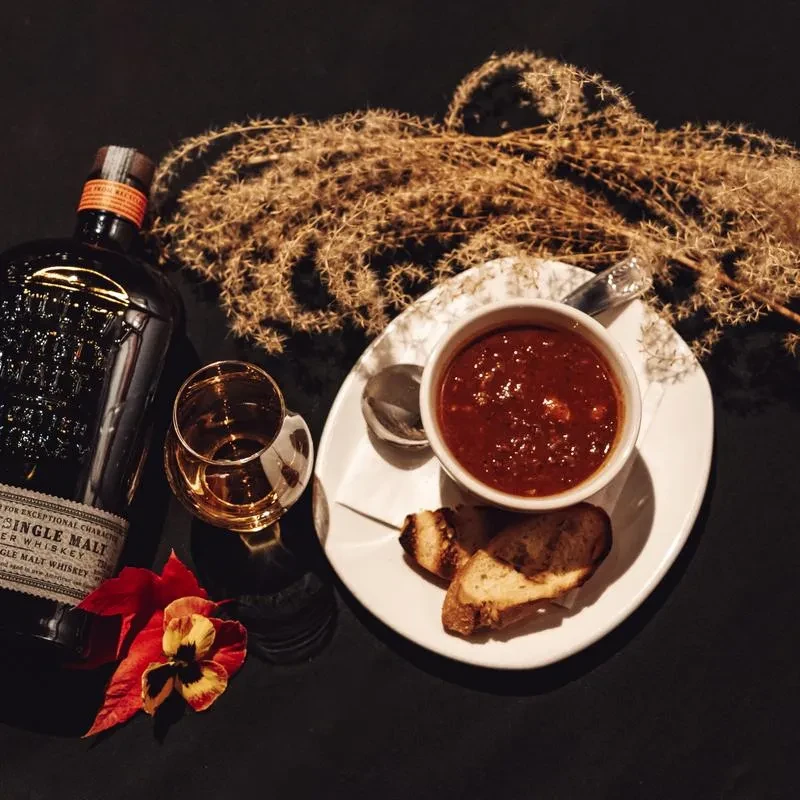 Top-down: whiskey, stew, red leaf, and dried plants on dark background.