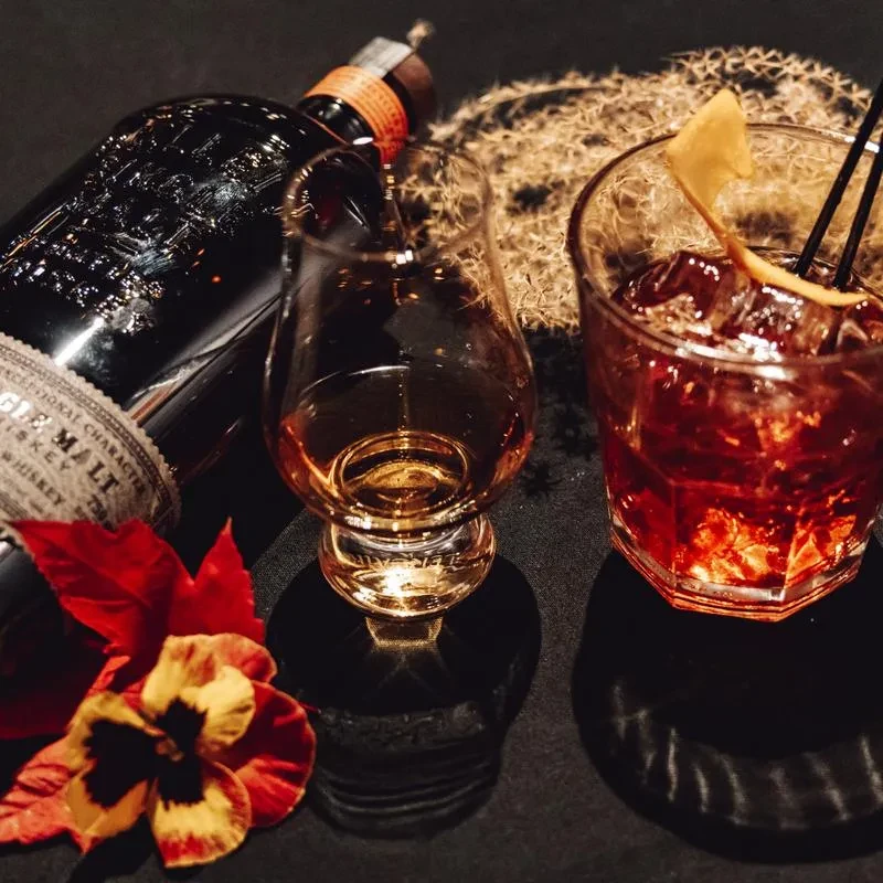 Whiskey bottle, tasting glass, and cocktail on a dark, moody background.