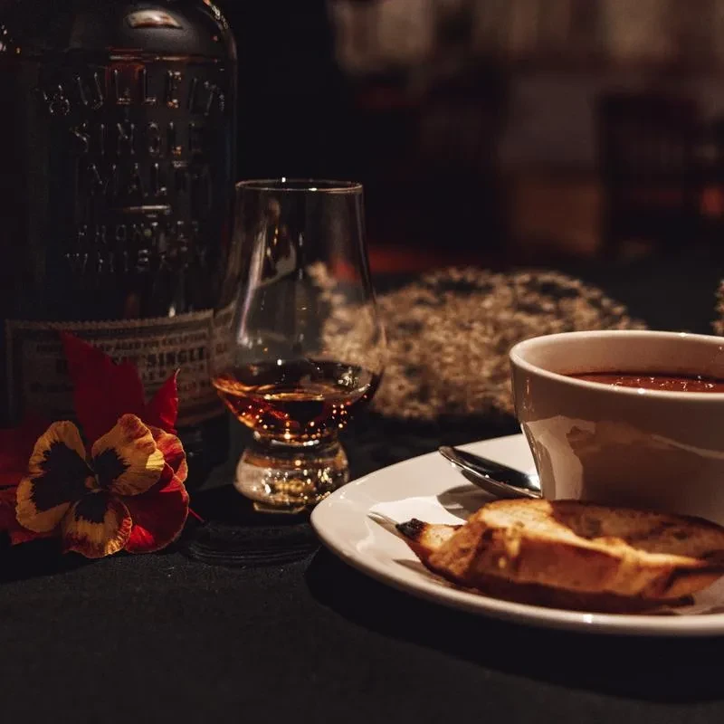 Warmly lit Bulleit whiskey bottle, snifter, cup, bread, and pansy flower.