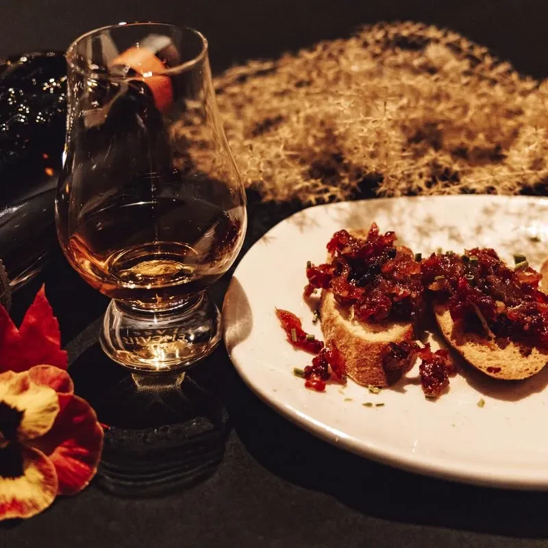 Dimly lit whiskey glass, bruschetta with relish, chives, and a flower.