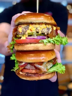 Close-up of hands holding two large, stacked burgers with cheese and bacon.