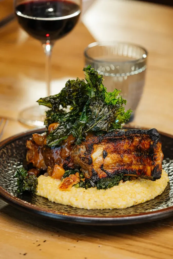 Roasted meat, creamy polenta, and crispy kale on a wooden table.