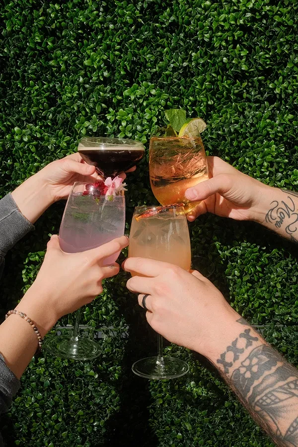 Four diverse hands raising distinct cocktails against a green hedge.
