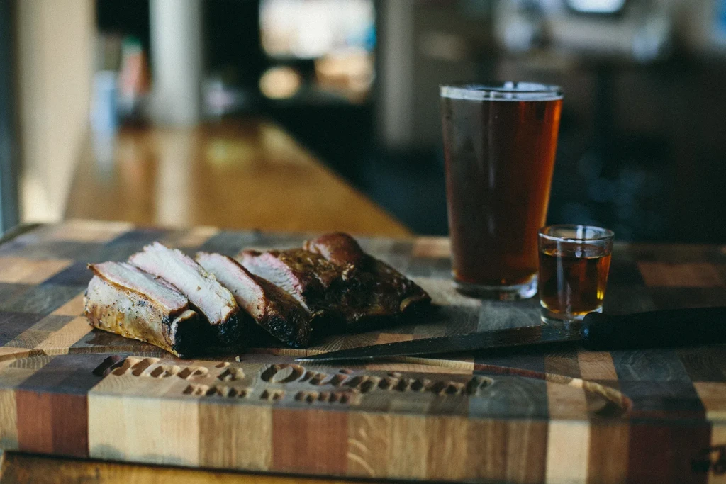 Sliced meat, beer, shot glass, and knife on a wooden cutting board.