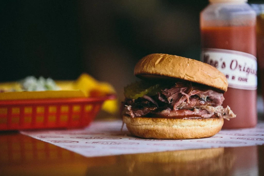 Close-up of a pulled pork sandwich, pickles, sauce, and BBQ bottle.