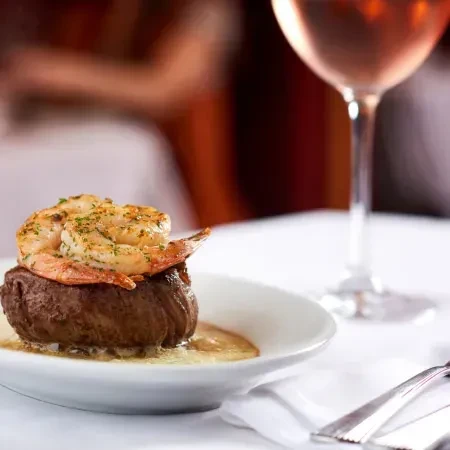 Thick steak topped with shrimp, rosé wine, and silverware in a restaurant.