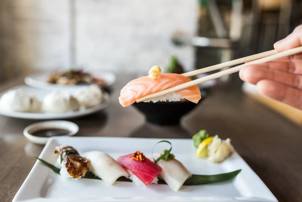Hand holding salmon nigiri with chopsticks over a plate of sushi.