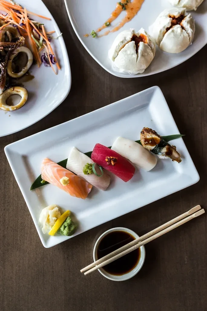 Overhead shot of sushi, calamari, and bao buns on a dark table.