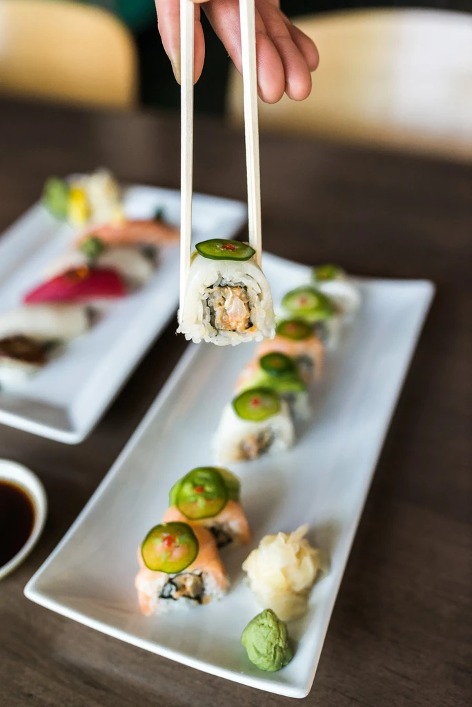 Chopsticks hold a sushi roll with green pepper over a sushi plate.
