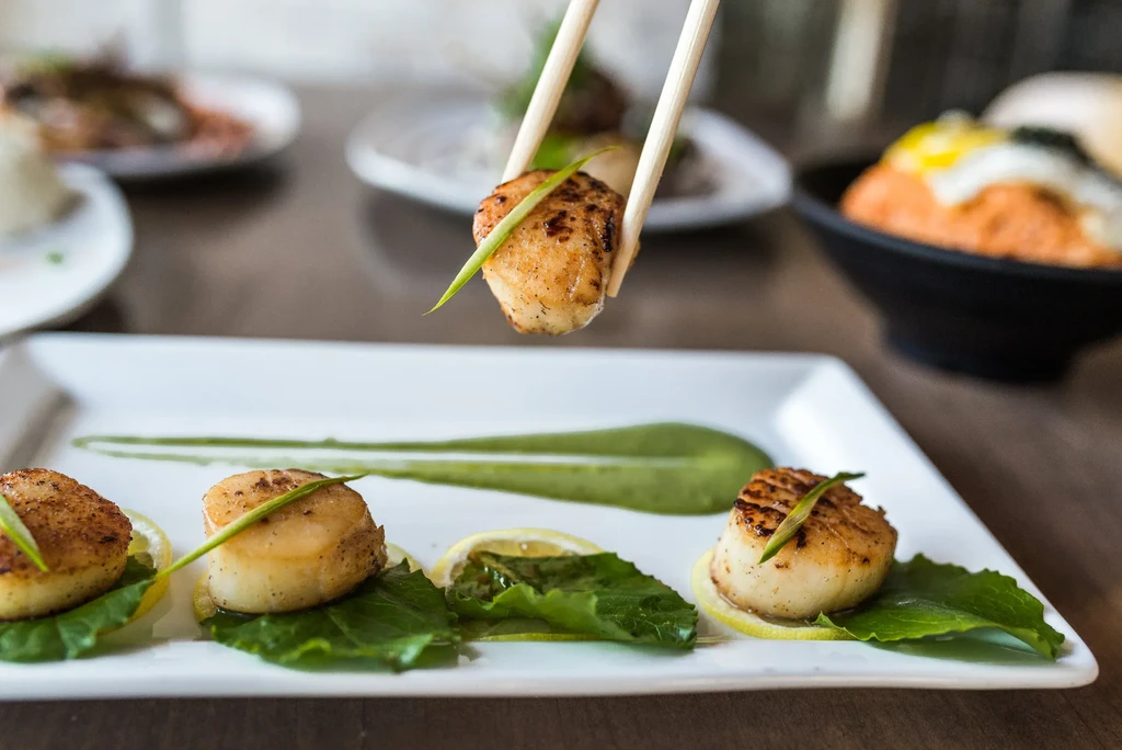 Pan-seared scallops with green sauce, one lifted by chopsticks.