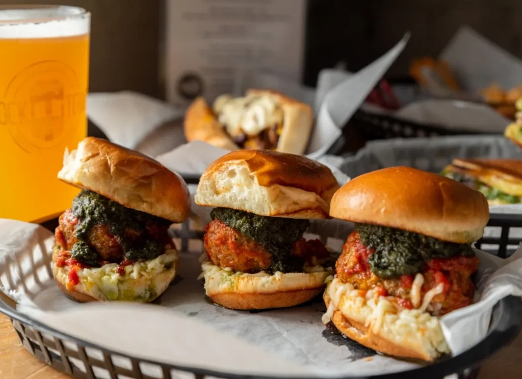 Three meatball sliders with red sauce, pesto, cheese, and beer.