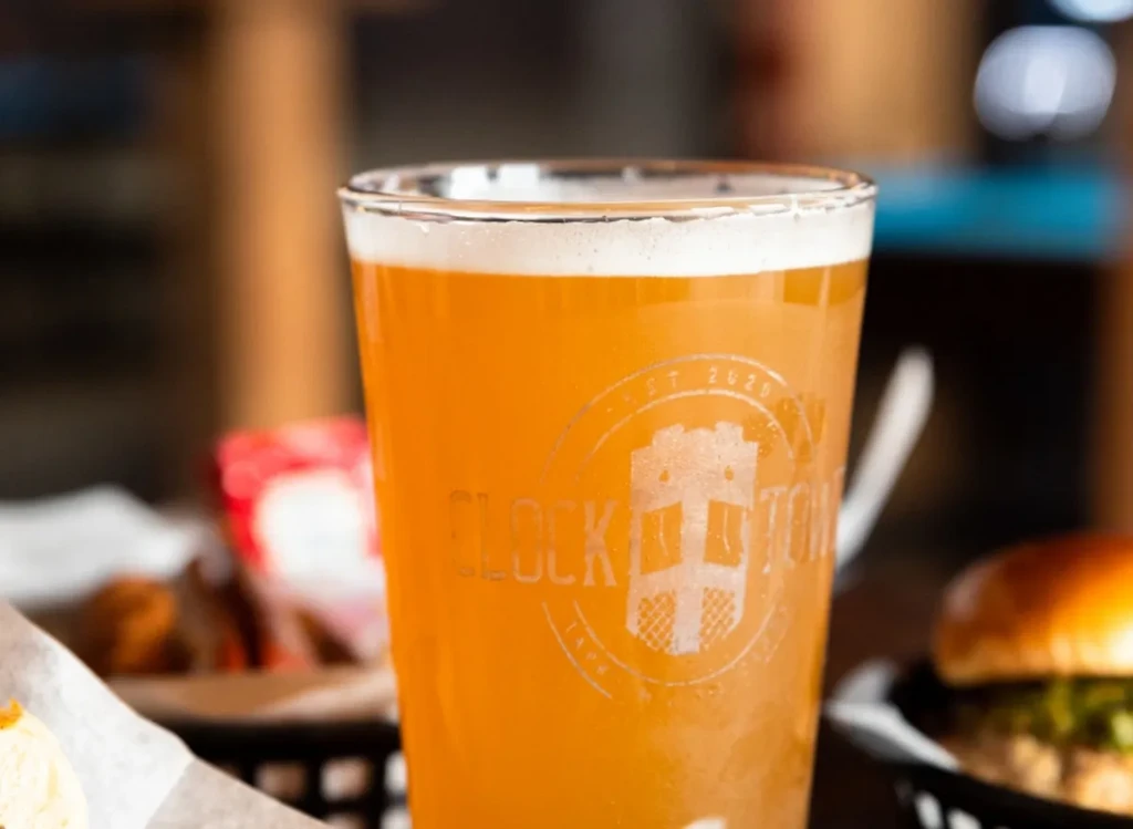 Amber beer in a Clock Town glass with foam and blurred food.
