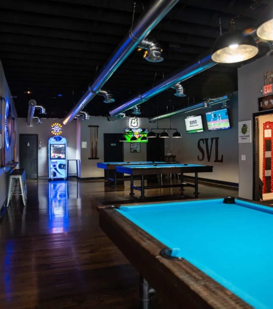 Modern pool hall interior with blue tables, industrial lights, and arcade games.