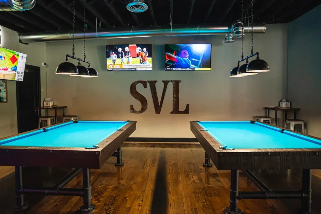 Industrial-chic game room featuring two turquoise pool tables and sports TVs.
