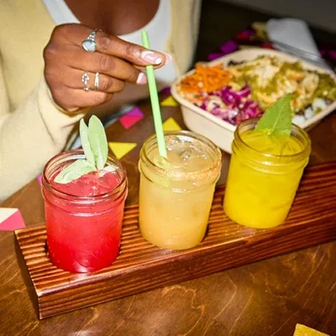 Three colorful mason jar drinks on a rustic tray, hand touching one.