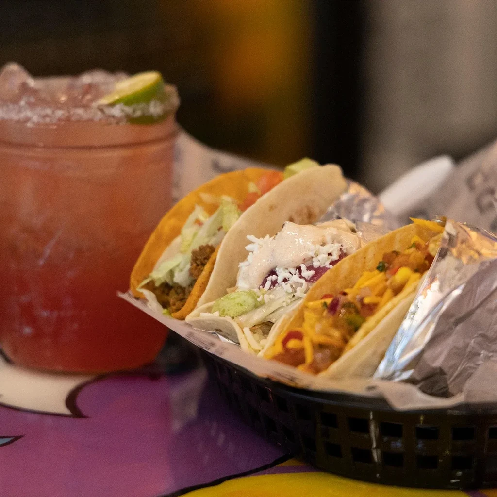 Four assorted tacos in a basket with a drink on a colorful table.