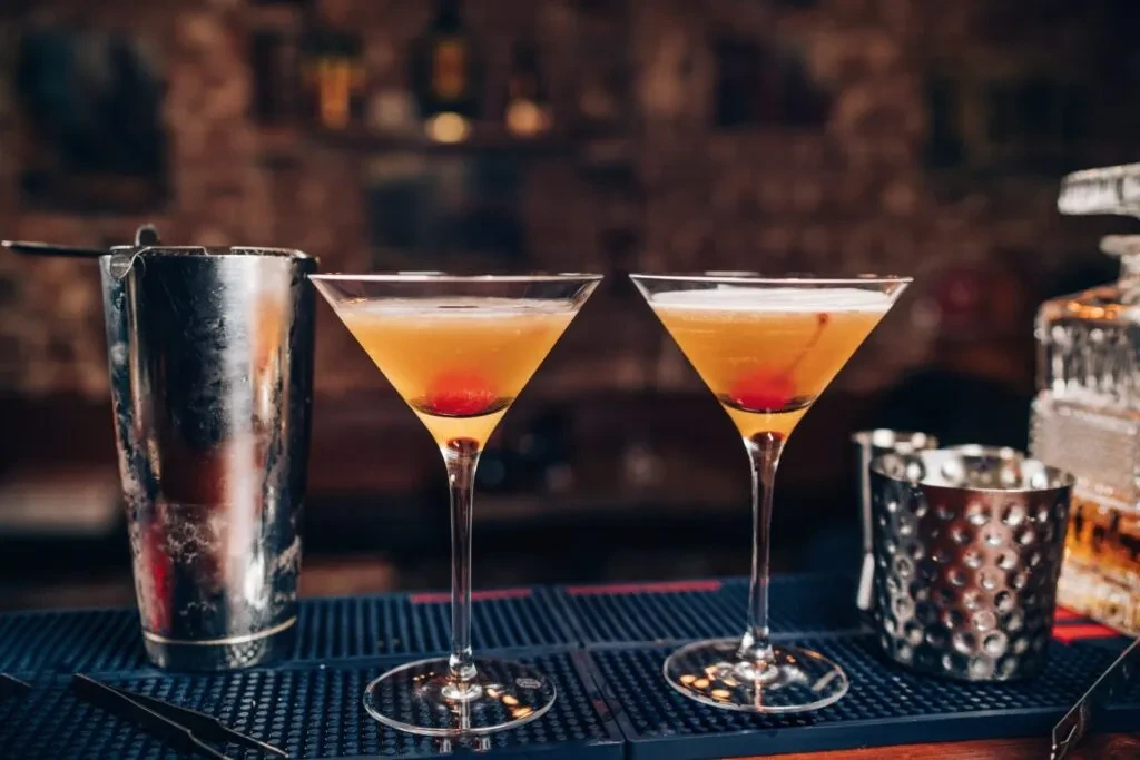 Two cherry cocktails, bar tools, and decanter on a dim bar.