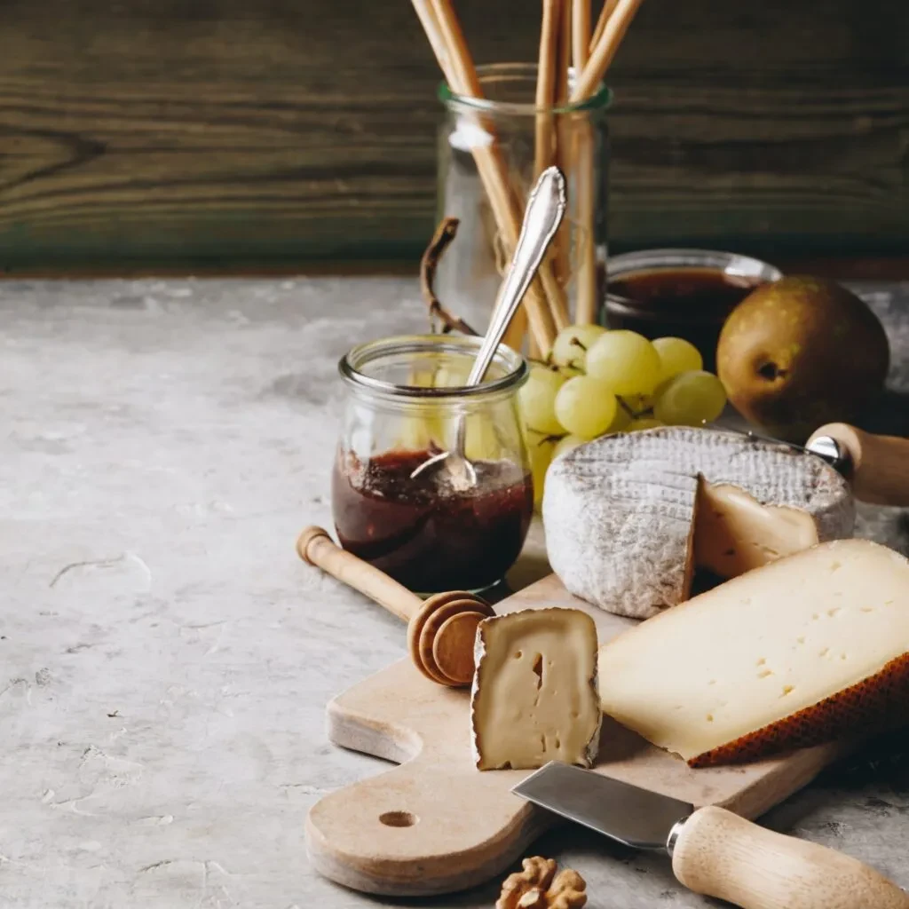 Rustic cheese board with various cheeses, jam, fruit, and breadsticks.