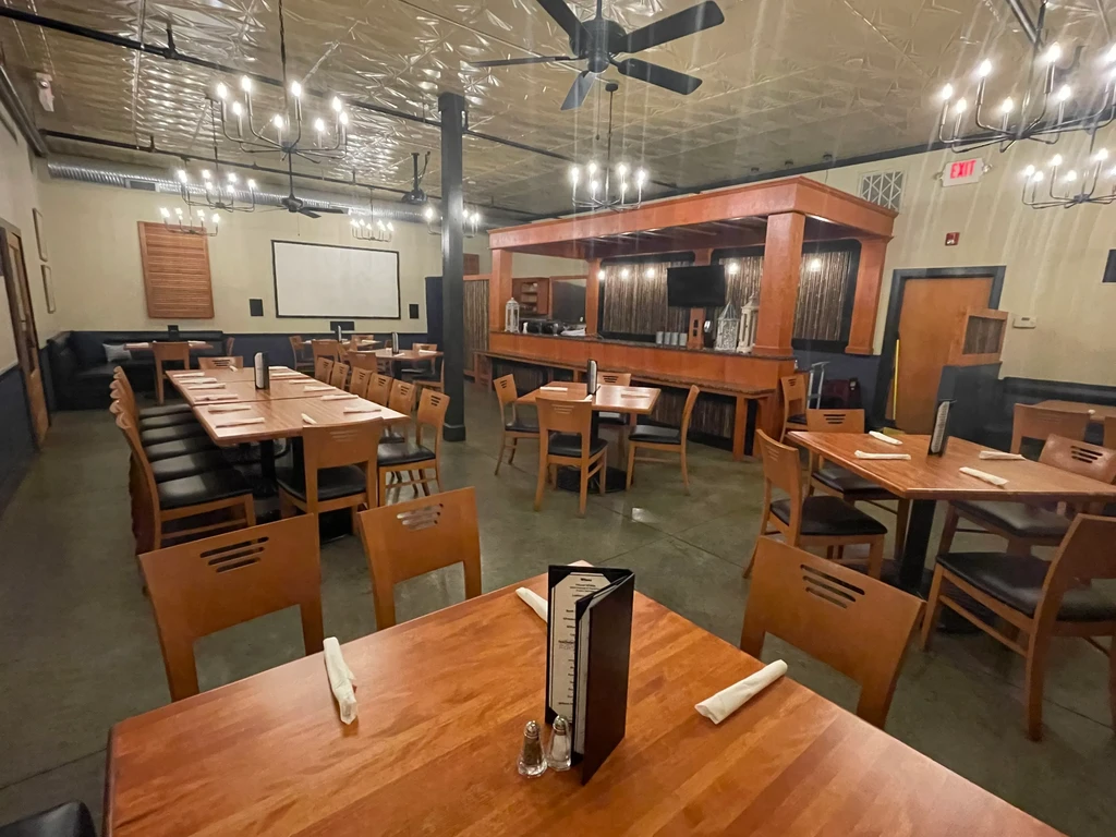 Empty restaurant interior: wooden tables, prominent bar, industrial lighting, screen.
