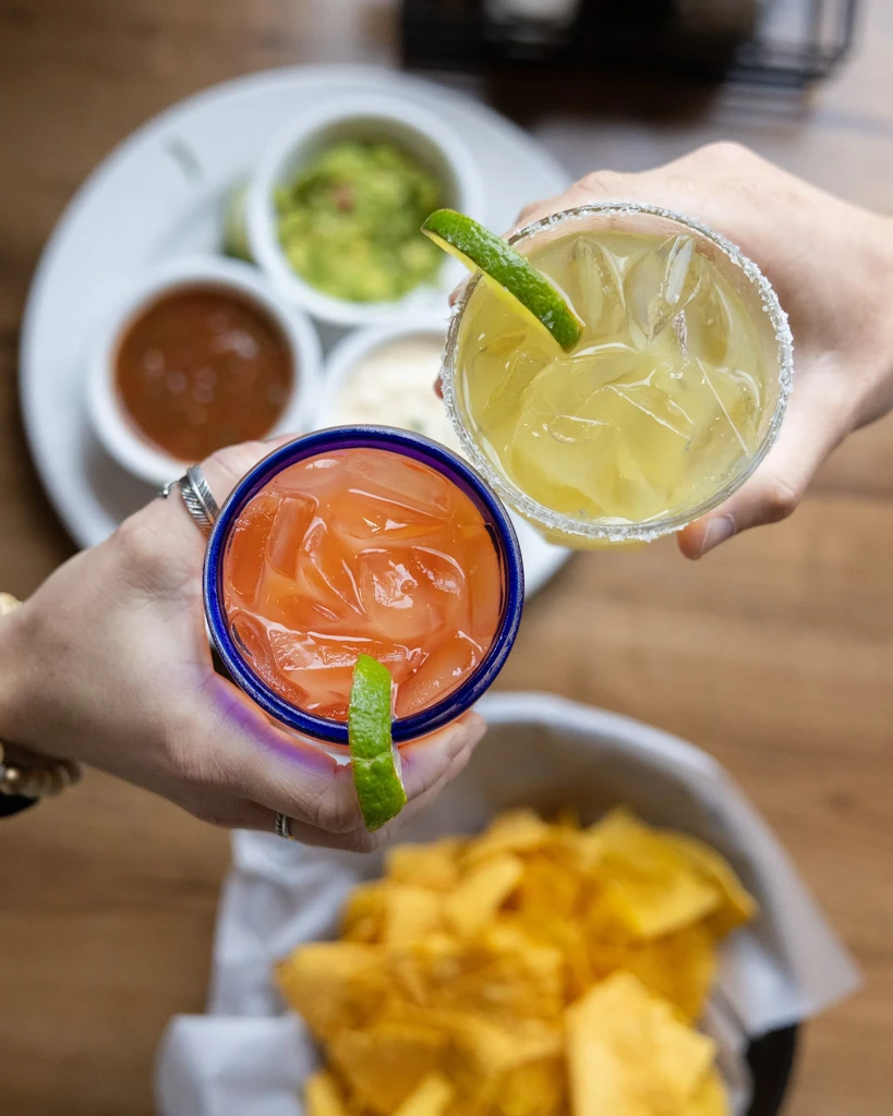 Two hands toast colorful cocktails garnished with lime, chips and dips.